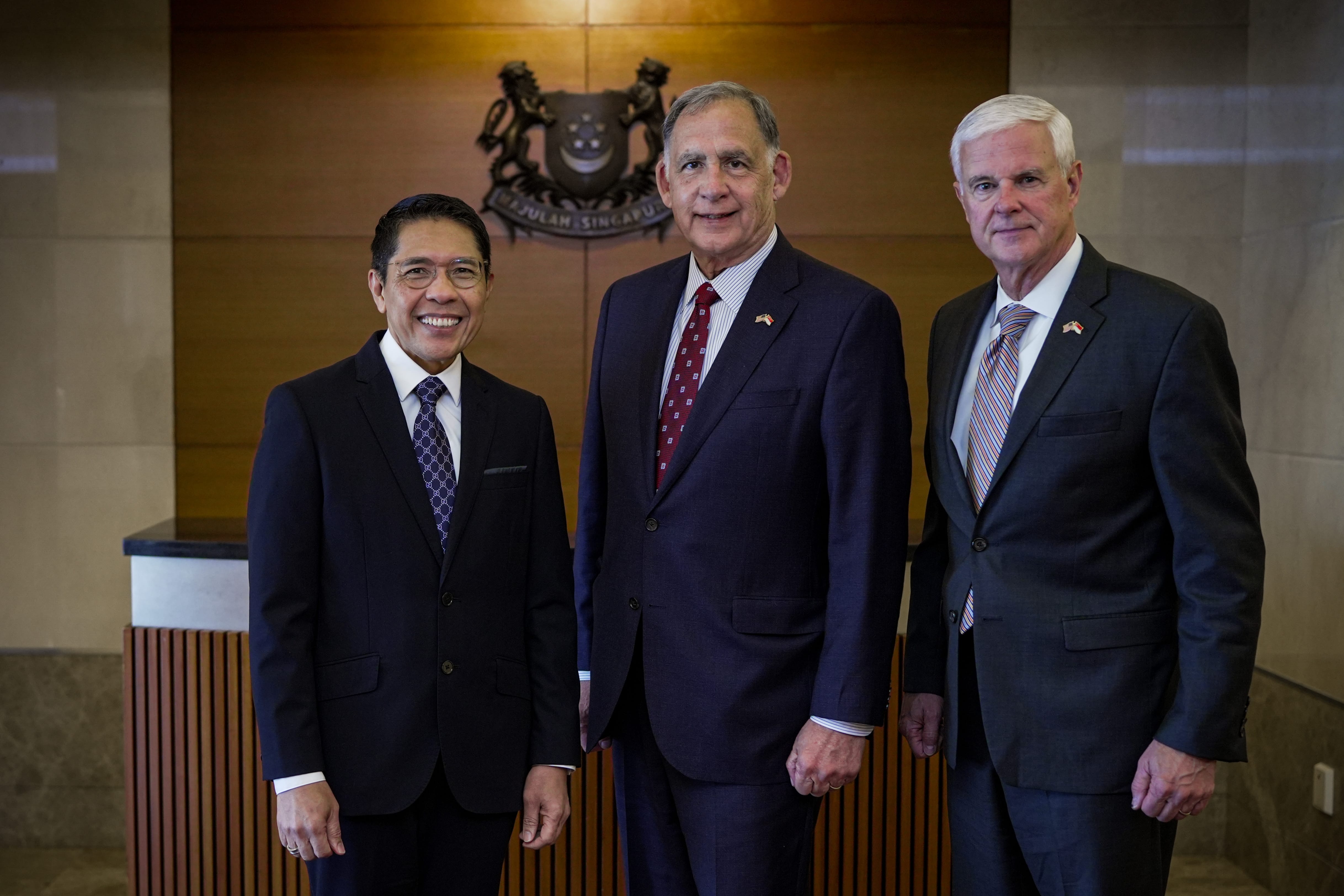 Three men in suits stand before a Singaporean crest on a wood paneled wall.